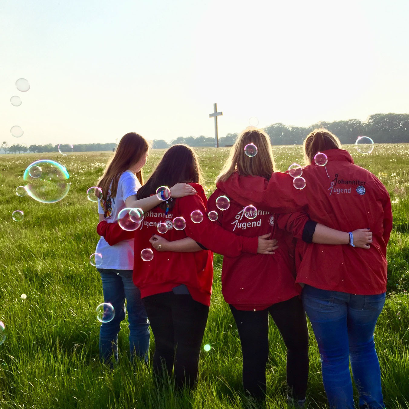 Vier Jugendliche mit Johanniter-Jugend-Jacke stehen Arm in Arm auf einer großen Wiese. Im Hintergrund ist ein großes Kreuz zu sehen.