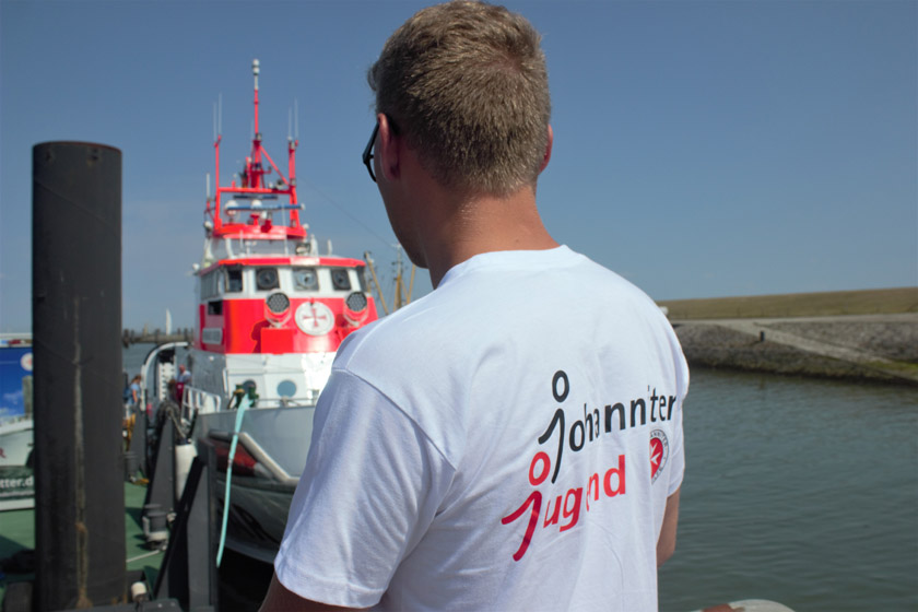 Ein Miglied der Johanniter-Jugend von ist von hinten zu sehen, er blickt auf ein im Hafen liegendes Boot.