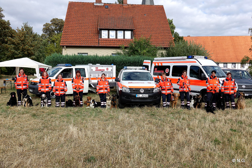 Gruppenbild Mitgliederinnen und Mitglieder der Rettungshundestaffel Südniedersachsen mit ihren Hunden vor den Johanniter-Einsatzfahrzeugen.