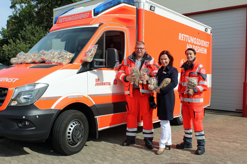 Johanniter gemeinsam mit Janet Hurst-Dittrich von der Sparkasse Hildesheim Goslar Peine und den Teddys vor einem Johanniter-Rettungswagen.