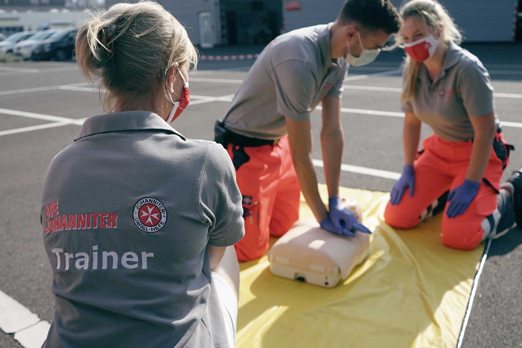 Erste-Hilfe-Training Eine Johanniter-Trainerin in grauem Poloshirt beobachtet zwei Personen, die an einer Puppe Wiederbelebung trainieren