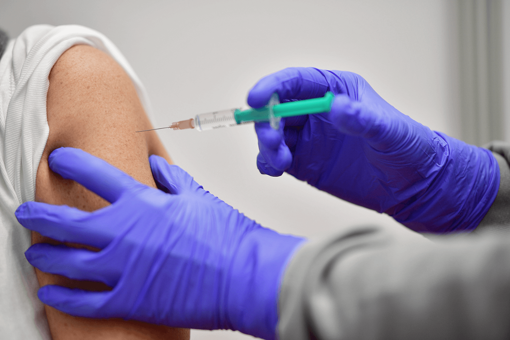 Corona-Impfung Der Fotoausschnitt zeigt zwei Hände in blauen Handschuhen, die eine Spritze in einen Oberarm geben.