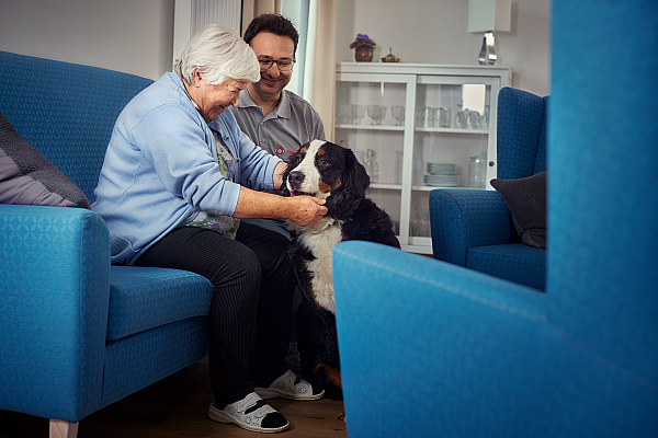 Ein Pfleger der Johanniter und eine Seniorin sitzen gemeinsam auf einem Sofa und sie streichelt einen Hund.