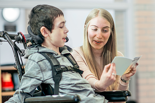 Eine Betreuerin spielt mit einem Jungen mit einer körperlichen Behinderung, der im Rollstuhl sitzt und lächelt.
