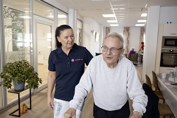 Eine pflegerin der Johanniter begleitet einen Tagesgast mit Rollator auf seinem Weg durch Tagespflegeflur