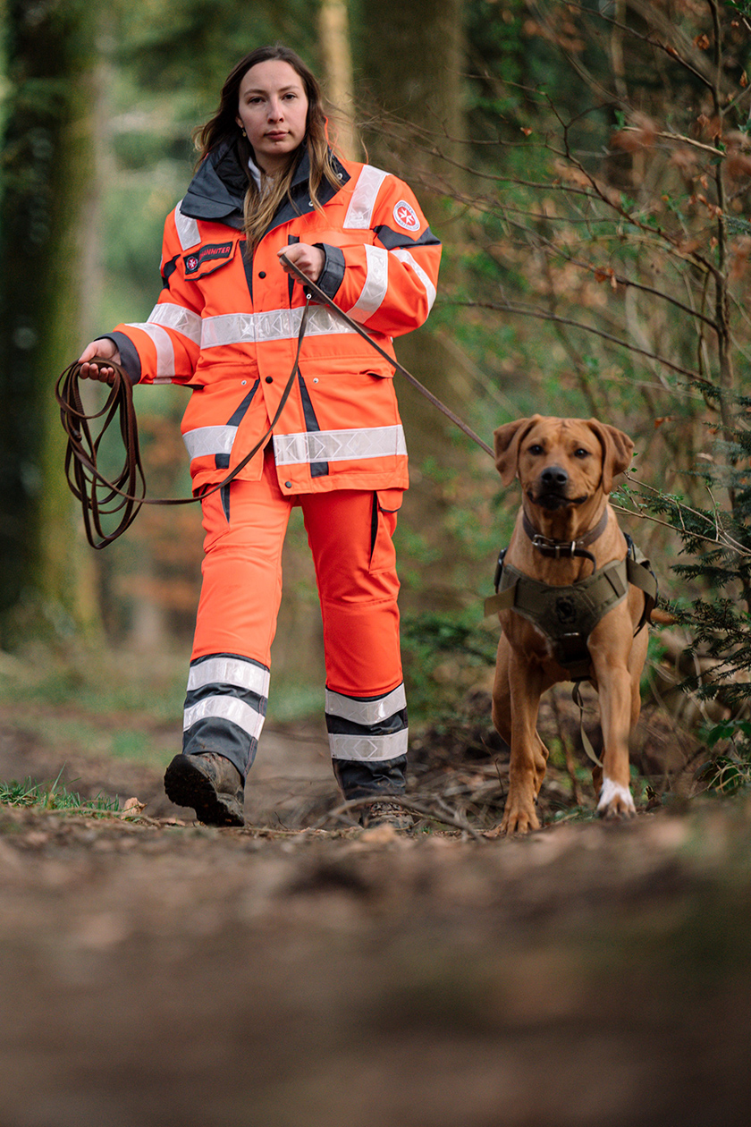 Suchhund an Leine mit Helferin in leuchtfarbener Einsatzkleidung.