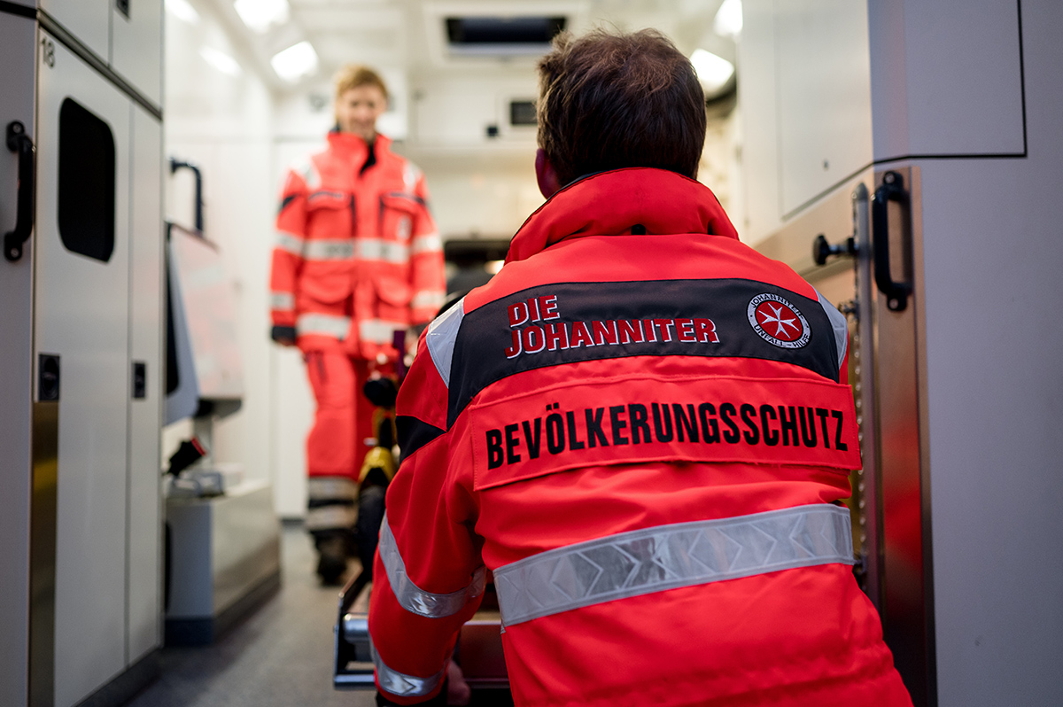 Eine Einsatzkraft des Bevölkerungsschutzes der Johanniter lädt eine Trage in einen Rettungswagen, während eine Kollegin im Hintergrund steht. Die auffällige rote Einsatzkleidung trägt die Aufschrift "Bevölkerungsschutz".