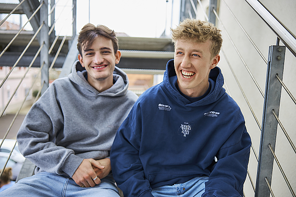 Zwei junge Männer, die lächelnd auf einer Treppe sitzen. Beide tragen Kapuzenpullover, einer in Grau und der andere in Dunkelblau. Die Stimmung ist entspannt und fröhlich.