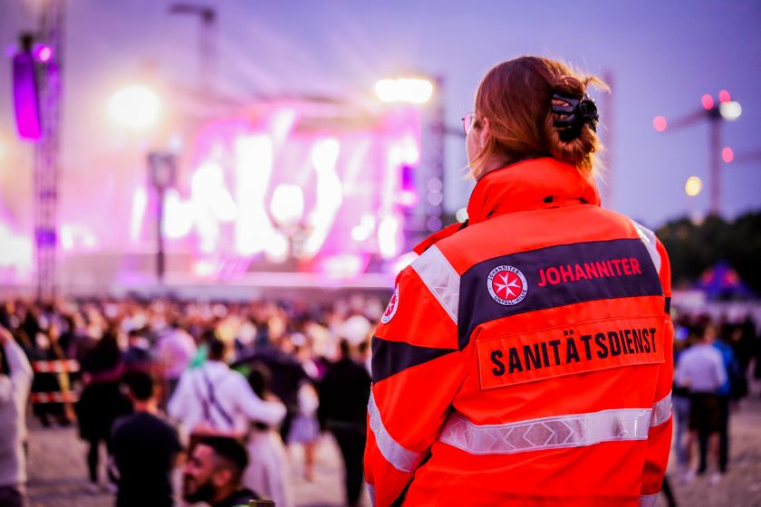 Eine Mitarbeiterin des Johanniter-Sanitätsdienstes steht mit dem Rücken zur Kamera vor einer großen Konzertbühne und überwacht die Menschenmenge auf dem Veranstaltungsgelände.