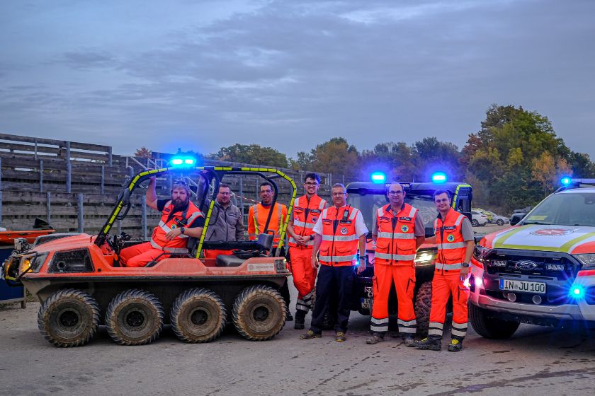 Johanniter-Einsatzkräfte in orangefarbener Schutzkleidung stehen neben Einsatzfahrzeugen mit eingeschaltetem Blaulicht.