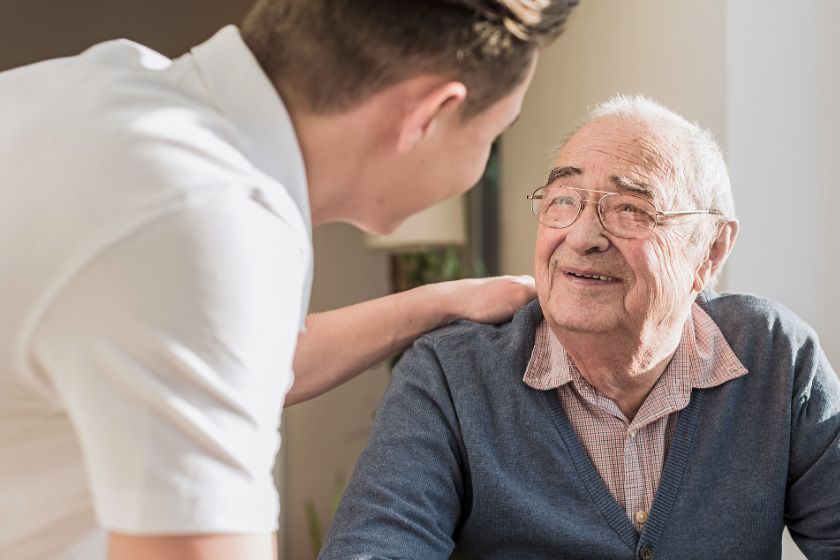 Wir sehen eine Pflegeperson, die einem älteren Mann freundschaftlich die Hand auf die Schulter legt.