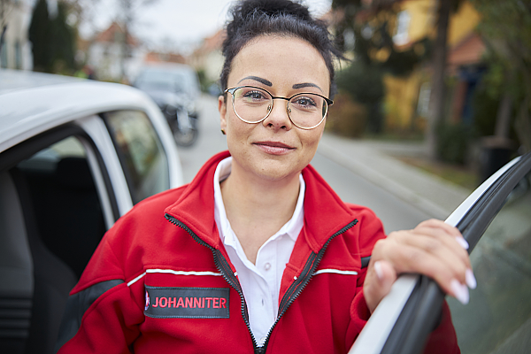 Eine Frau mit Brille und roter Johanniter Jacke steht in der Tür eines Fahrzeuges