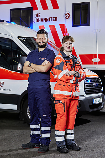 Zwei Freiwilligendienstleistenden stehen mit verschränkten Armen nebeneinander vor einem Einsatzwagen