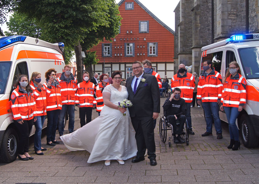 Lukas Hennemann und seine Frau stehen in Hochzeitskleidung neben zwei Einsatzfahrzeugen und den Johanniter die ihnen zur Vermählung gratulierten. 