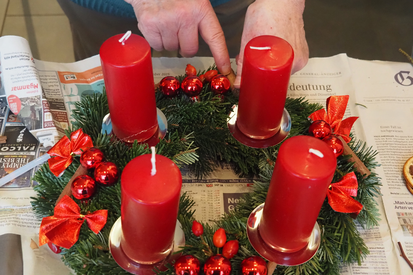 Heraus kommen wunderbare individuelle Kränze: hier ein Kranz aus Nobilis-Tanne, mit Schleifen und dicken Kerzen in rot gehalten. Dazu kleine rote Kugeln, die im Licht funkeln.