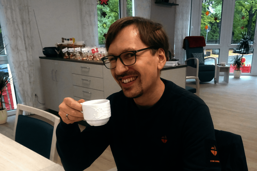 Ein dunkelblau gekleider Mann mit Brille hebt eine Kaffeetasse hoch und lächelt in die Kamera.