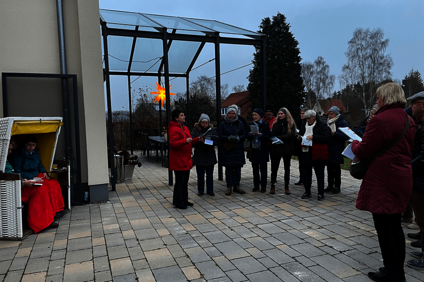 Eine Gruppe Sänger und Sängerinnen steht im Halbkreis auf einer Terrasse. Im Hintergrun dleuchtet ei Adventsstern; links sitzen zwei Personen zugedeckt in einem Strandkorb