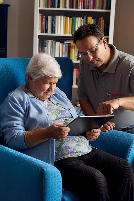 Zu sehen ist eine Frau, die in einem blauen Sessel sitzt und ein Tablet bedient, mit Unterstützung eines Pflegers