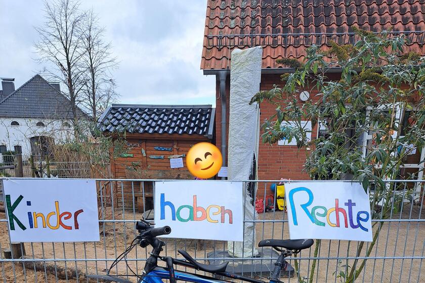 Am Zaun der Kita hängen drei selbst gemalte Plakate mit den Worten Kinder - haben - Rechte; im Hintergrund ist die Kita zu sehen.