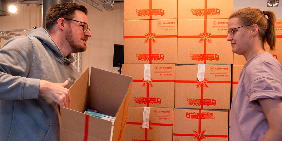 Ein hoher Stapel Weihnachtstrucker-Pakete davor ein Mann der einer Frau ein geöffnetes Paket reicht.