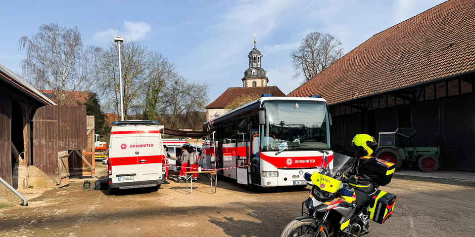 Ein Einsatzleitwagen und ein Bus mit Johanniter-Logo. Dazwischen Einsatzkräfte. Im Vordergrund steht ein Motorrad der Johanniter. Links und rechts sind Scheunen und im Hintergrund eine Dorfkirche zu sehen.