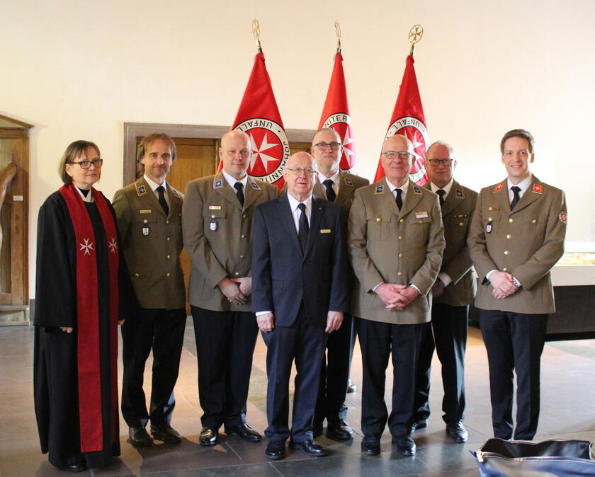 Eine Pfarrerin und 7 Männer, teils in Uniformen stehen vor 3 großen Johanniterfahnen