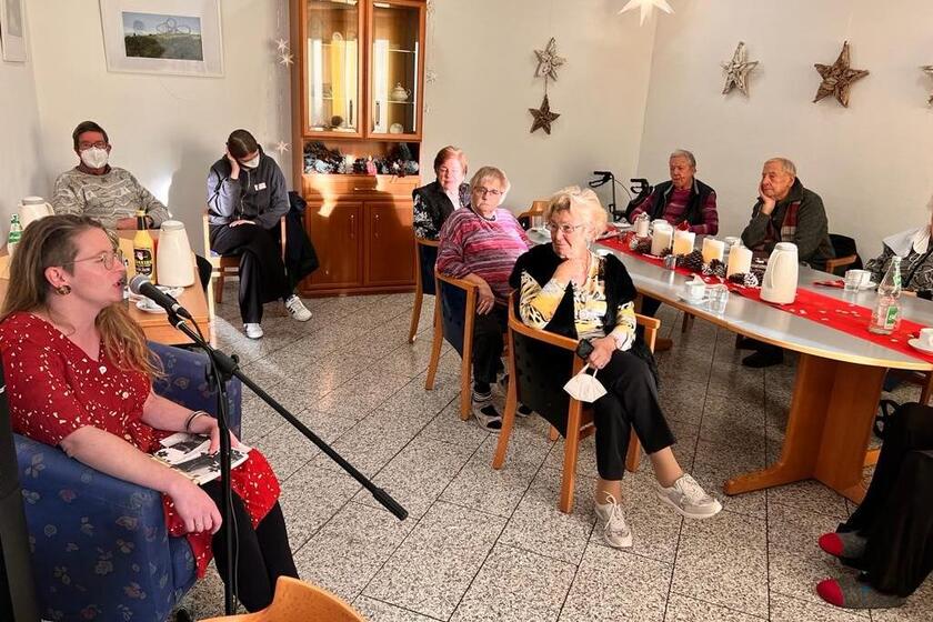 Eine Frau im roten Kleid sitzt vor einem Mikrofon; ringsherum sitzen Menschen und hören zu.