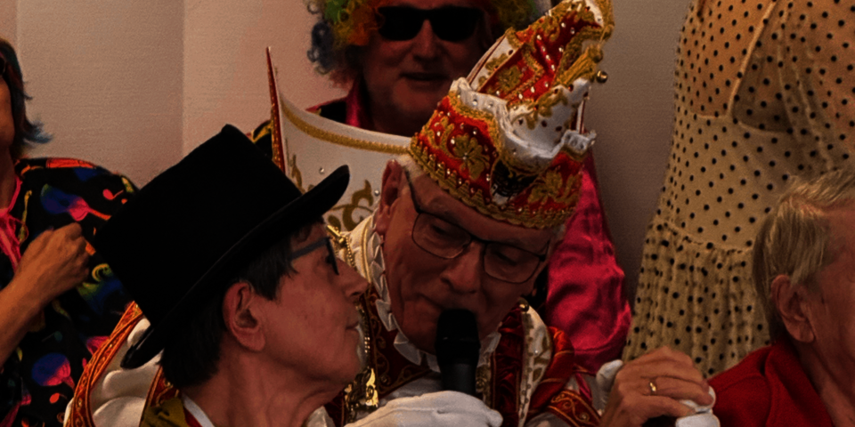 Mann mit Narrenkappe hält ein Mikrofon und spricht mit einem sitzenden Mann mit Hut