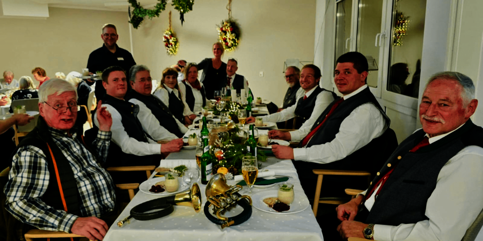 Lange festliche Tafel mit Jagdgesellschaft in Tracht, Menü und Getränken, grün geschmückt und weihnachtlich dekoriert.