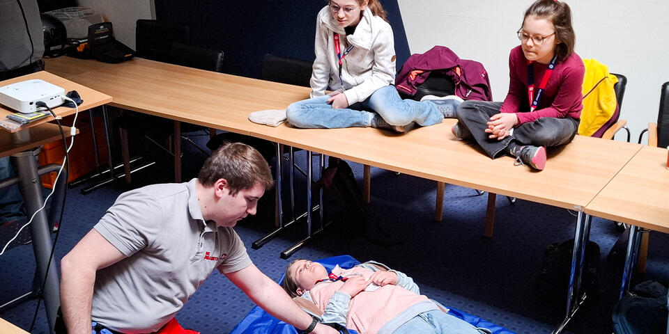 Ein junger Mann zeigt an einer Frau Erste-Hilfe-Maßnahmen. Kinder schauen zu.