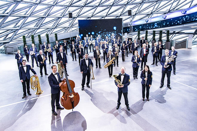 Gruppenfoto einer Orchesterkapelle in einem modernen, geometrischen Gebäude mit einer Glas-Stahl-Konstruktion. Die Musiker tragen Anzüge und Kleider, während sie ihre Instrumente präsentieren, darunter Tuben, Posaunen und Klarinetten.