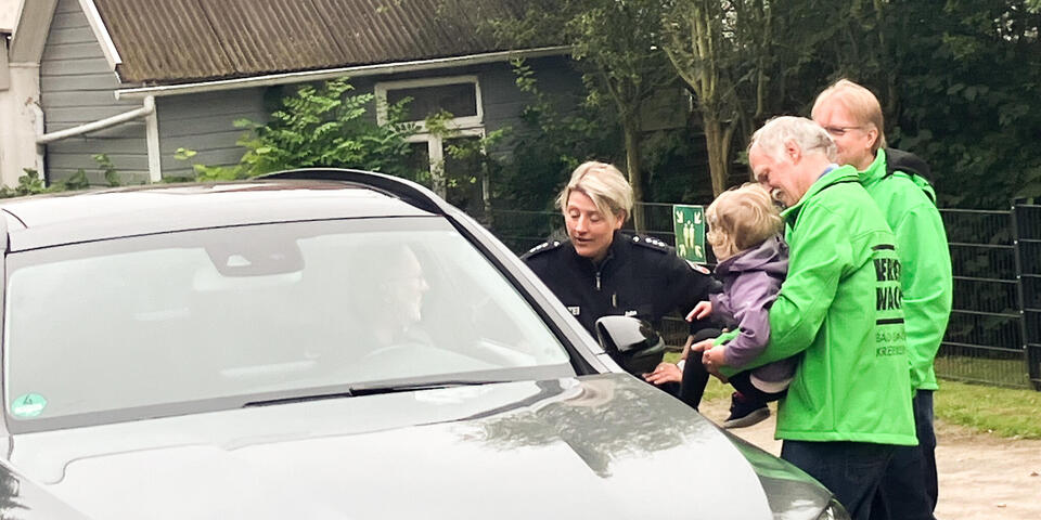 Zwei Männer, einer davon hält ein kleines Kind auf dem Arm und eine Polizistin sprechen mit einem Autofahrer