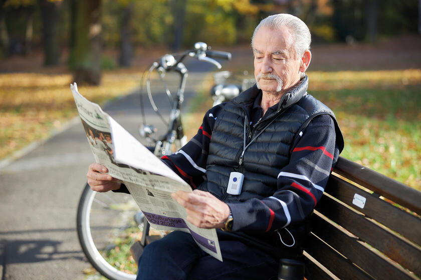 Ein Senior sitzt auf einer Parkbank und liest Zeitung, neben ihm steht sein Fahrrad, um den Hals hat er den Kombinotruf an einem Halsband hängen.