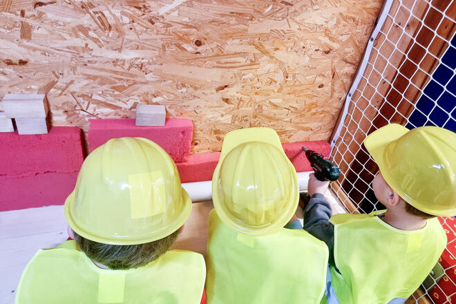 Drei Kinder mit Sicherheitswesten und Bauhelmen an einer Spanplatte