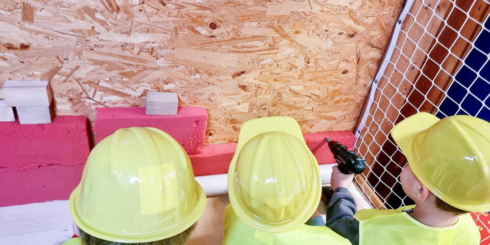 Drei Kinder mit Sicherheitswesten und Bauhelmen an einer Spanplatte