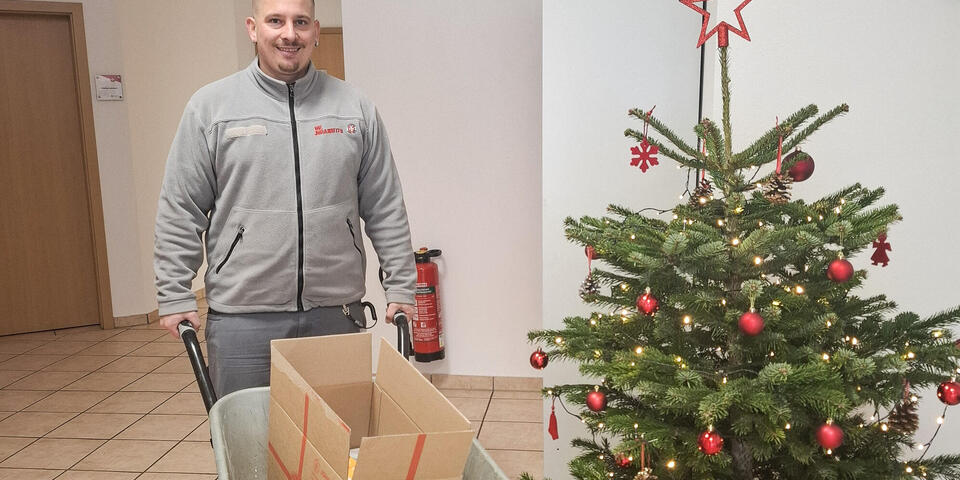 Ein Mann mit einer Schubkarre neben einem Weihnachtsbaum. In der Karre liegt ein geöffnetes Paket