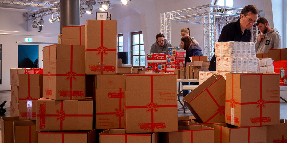 Im Vordergrund Weihnachtstrucker-Pakete. Dahinter packen Menschen weitere Pakete