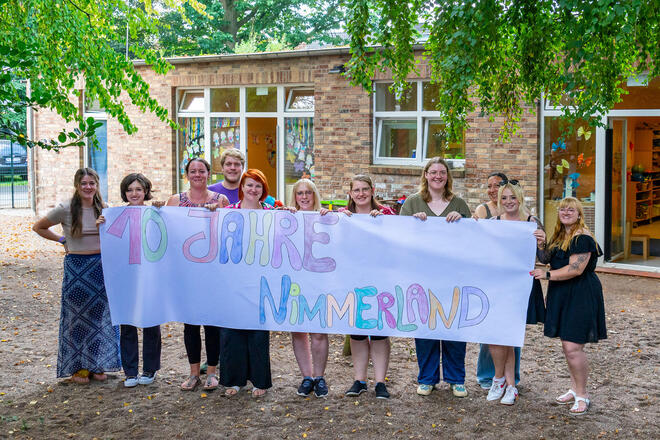 Das Team der Kita Nimmerland steht auf dem Spielplatz und hält ein Banner hoch, auf dem mit Buntstiften "10 Jahre Nimmerland" geschrieben steht.