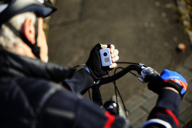 Blick über die Schulter eines Radfahrers auf den Link und eine Hand, die ein kleines weißes Kästchen mit einem Knopf in der Mitte hält