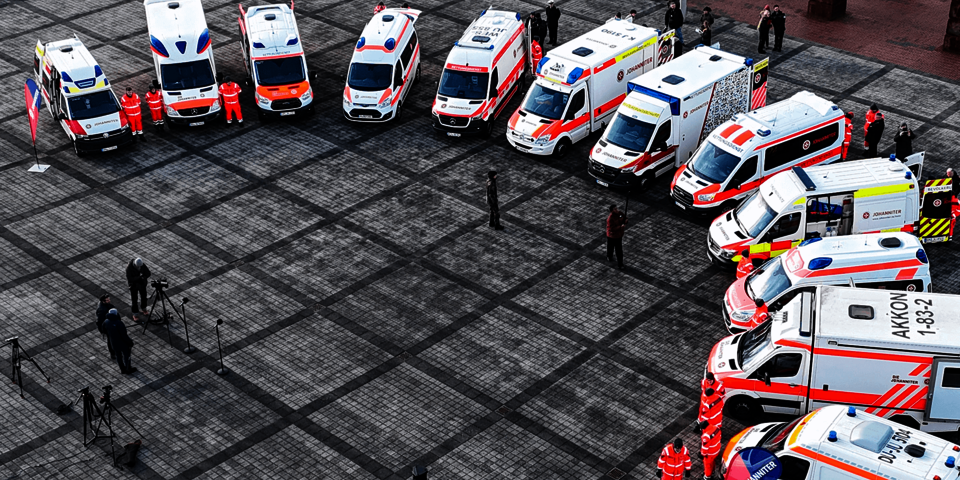 Vogelperspektive: Reihe von Rettungsfahrzeugen und Einsatzkräften auf einem großen gepflasterten Platz.