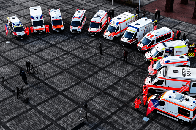 Vogelperspektive: Reihe von Rettungsfahrzeugen und Einsatzkräften auf einem großen gepflasterten Platz.