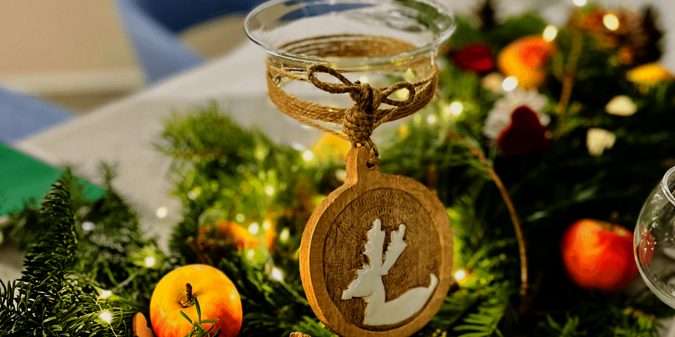 Nahaufnahme einer festlichen Tischdeko mit Tannenzweigen, gläsernem Windlicht, Holzanhänger mit Rentier-Motiv und Äpfeln.