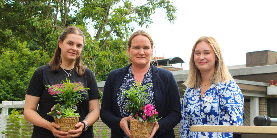 Zwei Frauen mit Blumen neben einer Frau im blauweißem Kleid.