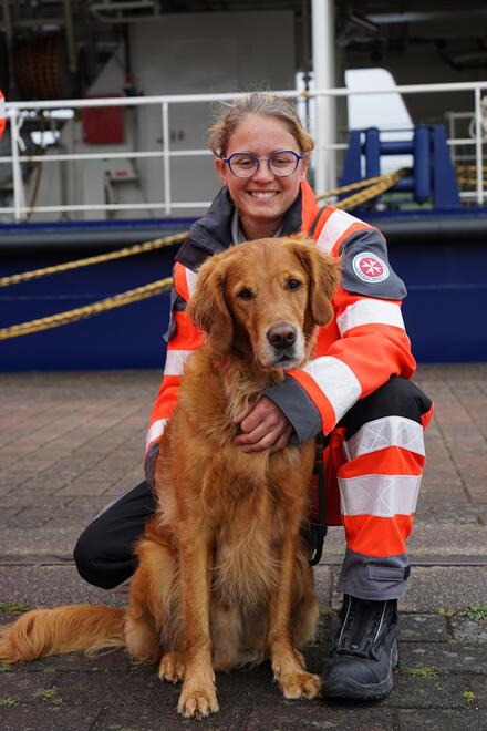Zu sehen ist eine Frau in Dienstkleidung, die einen Hund streichelt. Beide sitzen auf dem Boden.