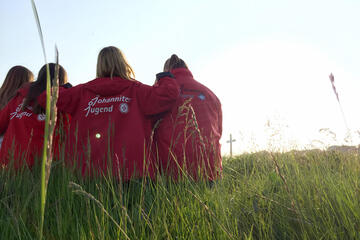 Vier Mädchen in Johanniter-Jugend-Kleidung sitzen auf einer Wiese und halten sich in den Armen. Im Hintergrund ist ein Kreuz zu sehen.