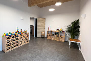 Heller Spielraum mit heller Echtholzdecke und dunklem Vinylboden in Holzoptik. Links Regale mit Schubladen für Bauklötze. Eine große Pflanze in der rechten hinteren Ecke.