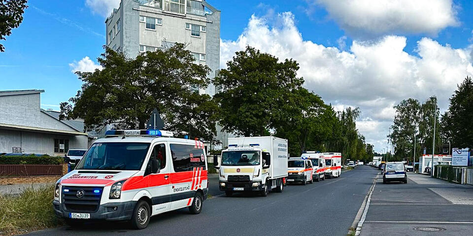 Johanniter-Einsatzfahrzeuge fahren auf einer Straße. Dahinter ein Hochaus