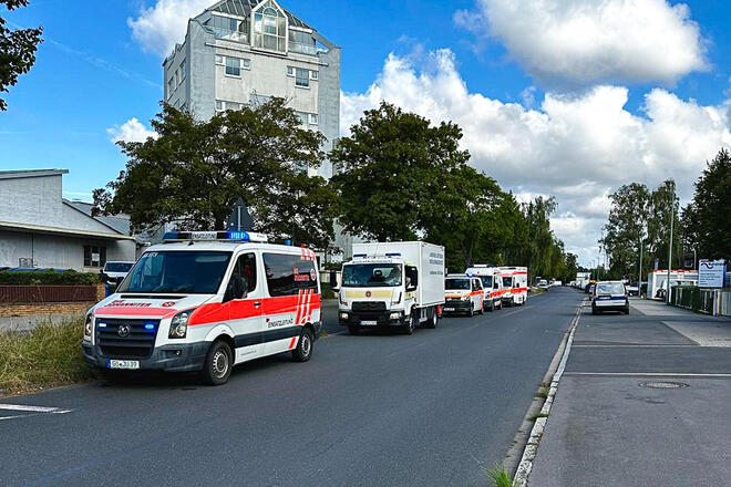 Johanniter-Einsatzfahrzeuge fahren auf einer Straße. Dahinter ein Hochaus