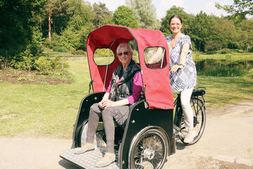 Eine ältere Frau wird von einer jüngeren Frau in einer Fahrrad-Rikscha durch einen Park gefahren