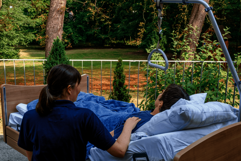 Ein Mensch mit kurzen dunklen Haaren liegt auf einer Terrasse in einem Bett und sieht in den Garten. Neben sitzt eine Frau in einem blauen Poloshirt und berührt seine Schulter.